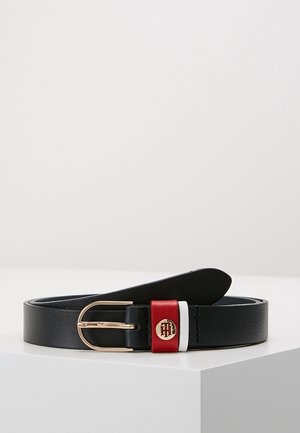 Zwarte leren riem met gouden gesp, met rode en witte leren lussen, en een rond gouden embleem op de rode lus, weergegeven op een witte ondergrond.