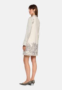 Beiges Kurzarmkleid mit einem Stadtmotiv am Saum. Der Stoff ist glatt und wird mit grauen spitzen Absatzschuhen kombiniert.