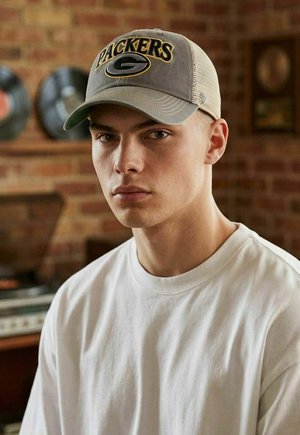 Junger Mann, der eine beige-graue Packers-Kappe und ein weißes Hemd trägt, mit einer Ziegelwand und Schallplatten im verschwommenen Hintergrund.