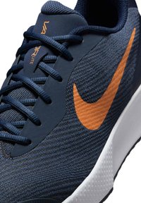 Baskets de sport bleu marine foncé avec une tige en maille texturée, accent de swoosh orange et semelle extérieure blanche robuste. Présente un design en couches et des lacets.