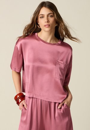 Femme aux cheveux lâches et ondulés portant un haut court à manches brillantes de couleur mauve et une jupe assortie, posant les mains dans les poches de la jupe sur fond neutre.