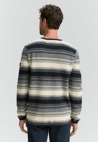 Man die met zijn rug naar de kijker staat, gekleed in een langemouwen sweater met horizontale zwarte, witte en grijze strepen.