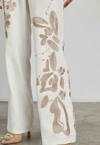 Pantaloni a gamba larga in tessuto bianco con delicate ricami floreali beige e motivi strutturati. Presentano una finitura liscia e una forma sartoriale.