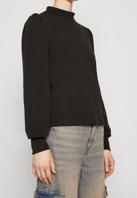 Schwarzes Langarmshirt mit hohem Kragen, mit gepufften Schultern und weichem Material. Kombiniert mit hellblauen Jeans.
