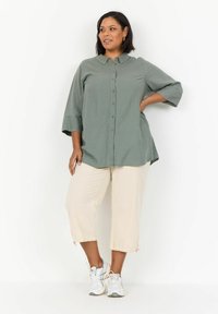 Green button-up shirt with three-quarter sleeves, paired with textured beige cropped pants and white sneakers. Simple and functional design.