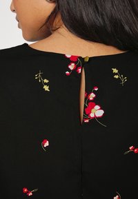 Kvinde set bagfra iført en sort blomsterkjole med en lille nøglehulåbning, der er fastgjort med en enkelt guldbutton ved halsen.