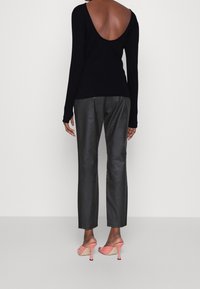 Woman wearing a black long-sleeve top with an open back, black straight-leg pants, and coral high-heeled sandals, standing against a plain background.