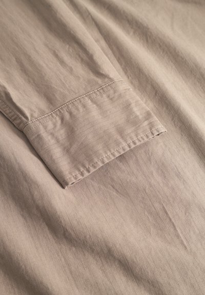 Tejido de camisa color beige claro con una textura suave, que presenta un puño cosido y un sutil patrón diagonal en toda su superficie.