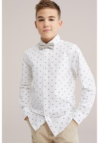 Rövid hajú fiú fehér mintás ingben, bézs csokornyakkendővel és bézs nadrágban, zsebeit tartva egy semleges háttér előtt áll.