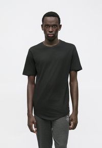 Jonge man die naar voren staat en een eenvoudige zwarte t-shirt en donkergrijze joggingbroek met een klein wit logo draagt.