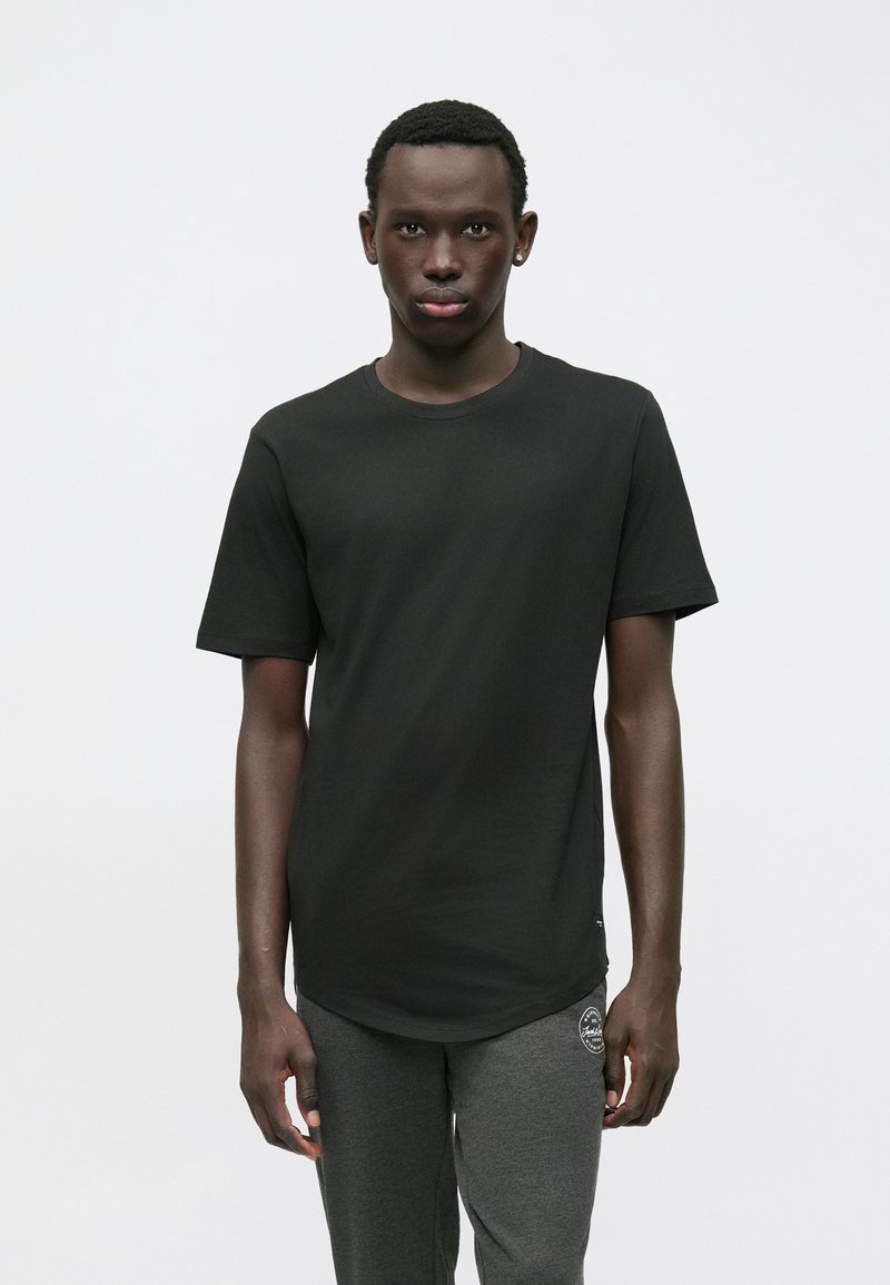 Jonge man die naar voren staat en een eenvoudige zwarte t-shirt en donkergrijze joggingbroek met een klein wit logo draagt.