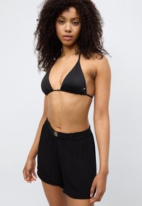 Μαύρο τοπ μπικίνι με τριγωνικά κύπελλα και λεπτούς ιμάντες, συνδυασμένο με άνετο μαύρο σορτς που έχει χρυσή λεπτομέρεια στη μέση.