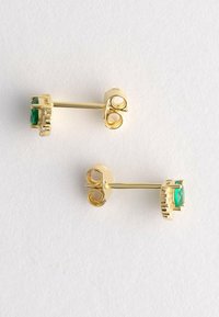 Orecchini a perno dorati con piccoli gemme verdi e chiusure a farfalla su una superficie bianca e texturizzata.