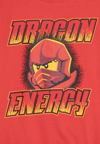 Piros póló, amelyen egy maszkos karakter grafikai terve látható. A felirat sárga és fekete vastag betűkkel olvasható, narancssárga akcentusokkal: "DRAGON ENERGY".