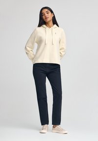 Krämfärgad huvtröja med dragsko, tillsammans med svarta byxor och ljusbeige sneakers. Avslappnad passform, mjukt tyg, minimal logotypdetalj på huvtröjan.