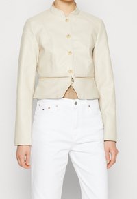 Veste en cuir crème raccourcie avec un col montant, des boutons dorés et une fermeture éclair asymétrique à la taille. Portée sur un jean blanc.
