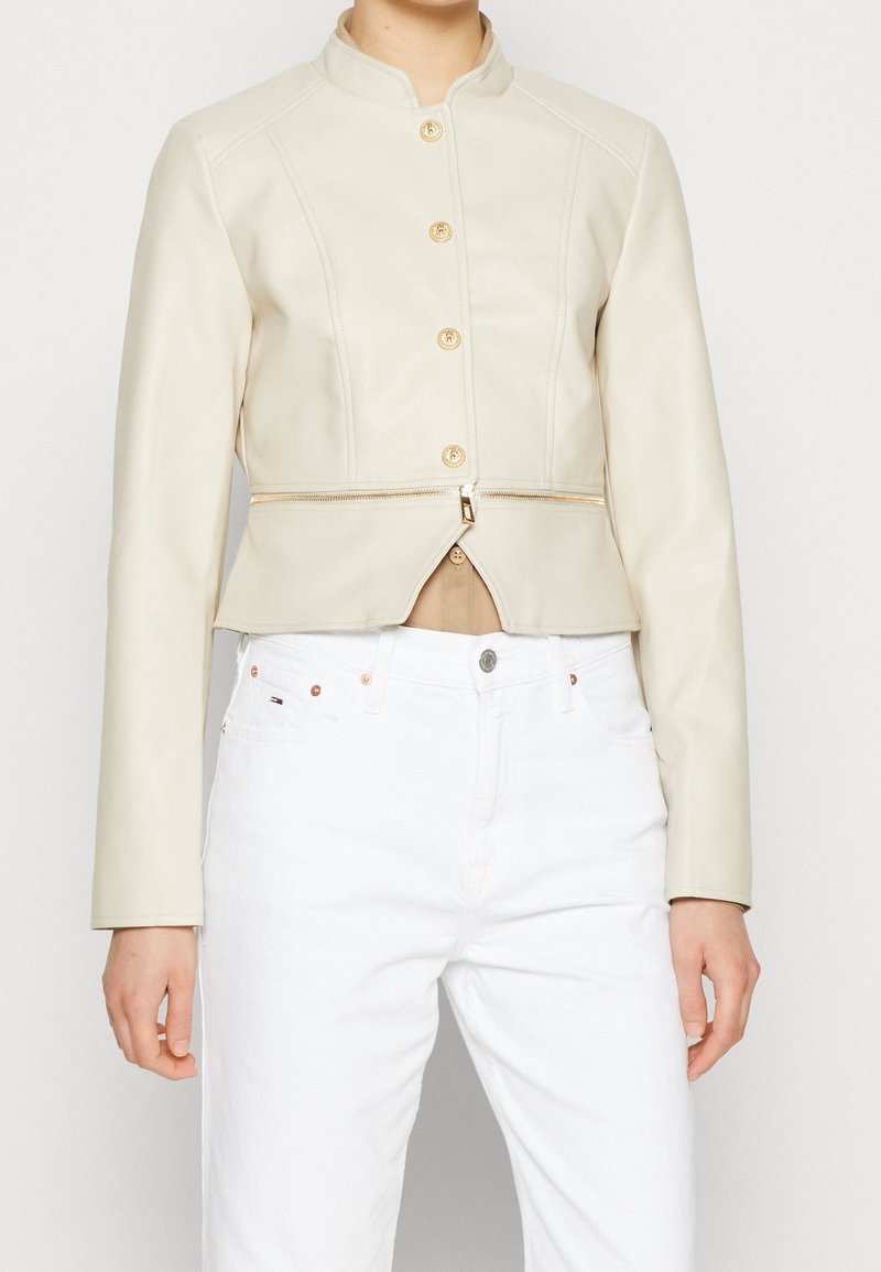 Veste en cuir crème raccourcie avec un col montant, des boutons dorés et une fermeture éclair asymétrique à la taille. Portée sur un jean blanc.