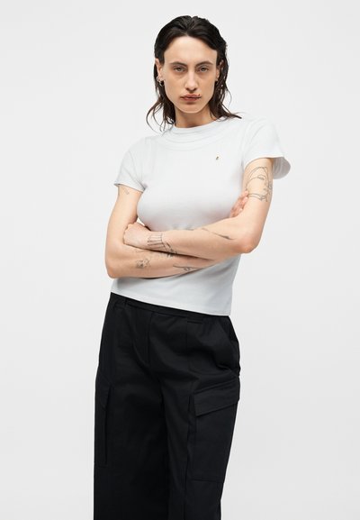 Personne aux cheveux foncés, T-shirt blanc moulant, pantalons cargo noirs, bras croisés et tatouages visibles sur les deux avant-bras et le haut du bras.