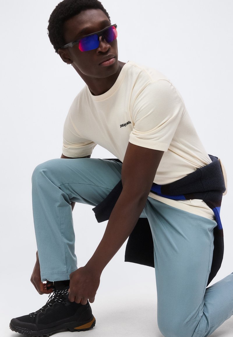 Mann mit reflektierenden Sonnenbrillen, beigem Patagonia-T-Shirt, hellblauer Hose, schwarzen Wanderschuhen, kniet und bindet seinen Schuh, mit einer dunklen Jacke um die Taille gebunden.