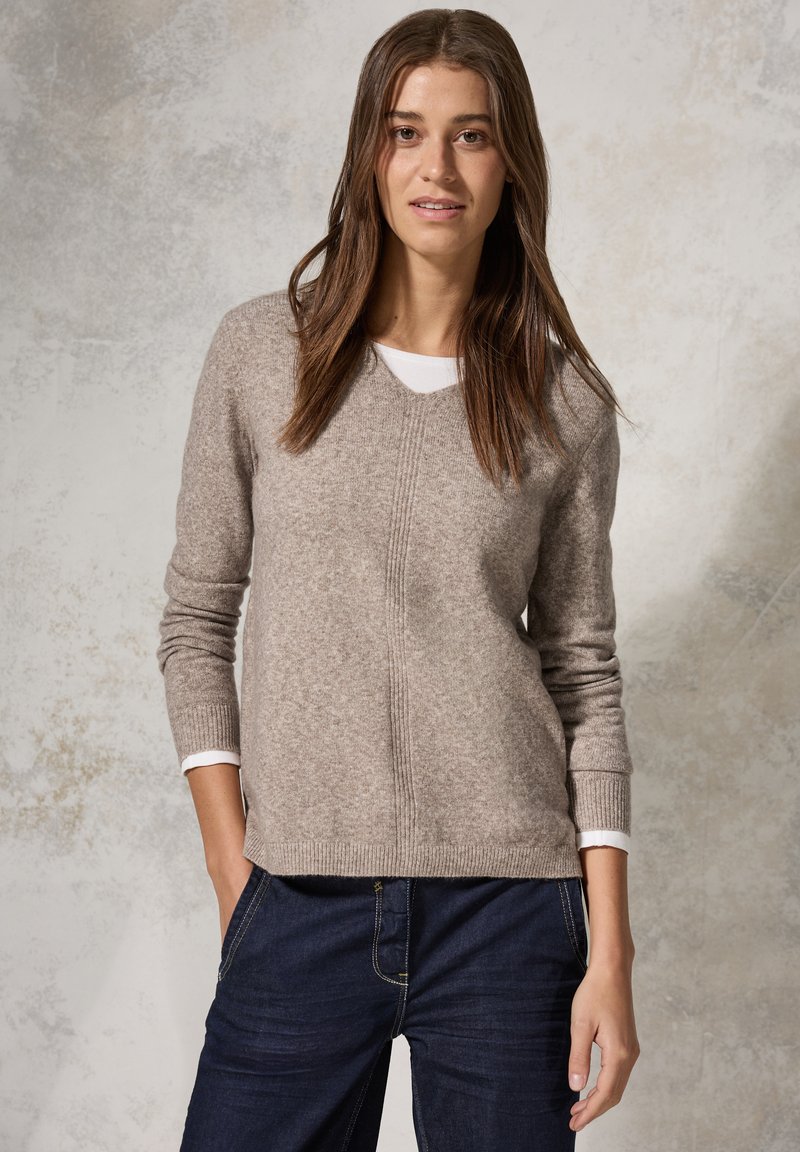 Maglione in lana marrone chiaro con scollo a V, polsini a costine e una finitura texturizzata, abbinato a jeans in denim blu scuro e una camicia bianca sotto.