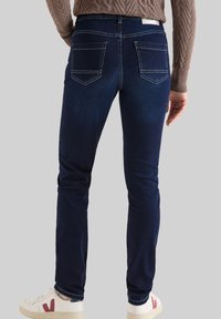 Jeans de color azul oscuro con un corte ajustado, fabricados en denim. Presentan dos bolsillos traseros con costuras en contraste y una cinturilla alta. Combinados con zapatillas blancas.