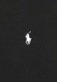 Logo brodé blanc représentant un joueur de polo à cheval tenant un maillet, sur un fond de tissu noir.