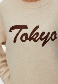 Beige gebreide trui met "Tokyo" in grote donkerbruine schreefletters geborduurd over de borst.