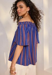 Top a righe blu e viola, senza spalline, con dettagli elastici sul collo e maniche corte, abbinato a pantaloni chiari.