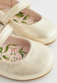 Zapatos Mary Jane de imitación cuero dorado con superficie texturizada, puntera redonda y plantilla con estampado floral. Cuenta con una sola correa con hebilla.