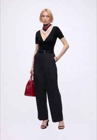Femme aux cheveux blonds courts portant un haut noir à col en V profond, un pantalon noir taille haute, un foulard rouge autour du cou, des talons noirs, tenant un sac à main rouge.