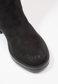 Bottine en daim noir à bout arrondi et semelle en caoutchouc sur fond blanc.