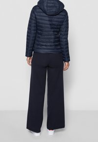 Marinblå pufferjacka med horisontell quiltning och huva, i kombination med vida marinblå byxor och vita sneakers, sedd bakifrån.
