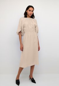 Frau in einem beigen Midikleid mit gerafft Taille und Puffärmeln, kombiniert mit schwarzen spitzen Slingback-Absätzen, die vor einem schlichten Hintergrund posiert.