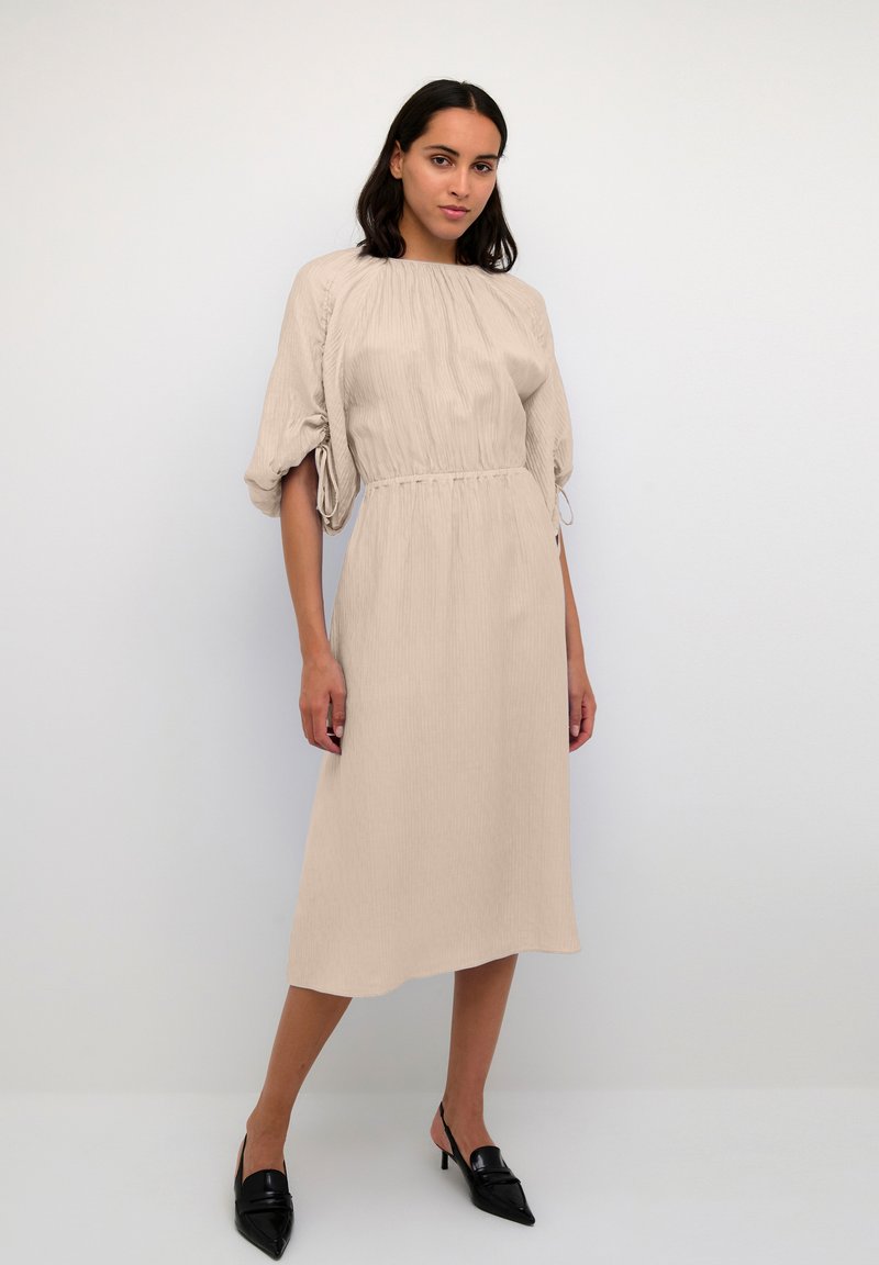 Frau in einem beigen Midikleid mit gerafft Taille und Puffärmeln, kombiniert mit schwarzen spitzen Slingback-Absätzen, die vor einem schlichten Hintergrund posiert.