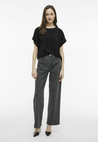 Juoda per didelė t-shirt su trumpomis rankovėmis, dėvima su aukštu liemeniu plačiomis pilkomis džinsomis ir juodais smailiais aukštakulniais, stovint prieš baltą foną.