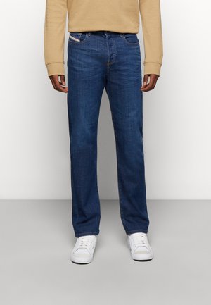 Pantalones vaqueros oscuros de corte recto, hechos de denim, con un bolsillo frontal y pasadores para cinturón. Combinados con zapatillas blancas y un sudadera beige.