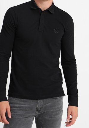 Czarna koszulka polo z długim rękawem, kołnierzykiem, dopasowanym krojem, teksturowaną tkaniną i małym emblematem na piersi. Połączona z szarymi jeansami.