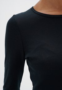 Juodas ilgas rankovėmis topas, pagamintas iš raukšlėtos medžiagos, su apvalia iškirpte ir prigludusia forma, akcentuojantis lygų paviršių ir paprastą dizainą.