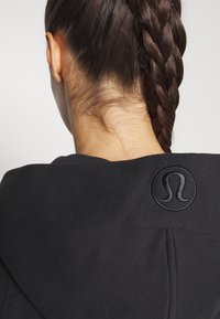 Czarna bluza z kapturem o gładkiej fakturze i okrągłym, wytłoczonym logo na plecach. Włosy są zaplecione w warkocz, eksponując dekolt.