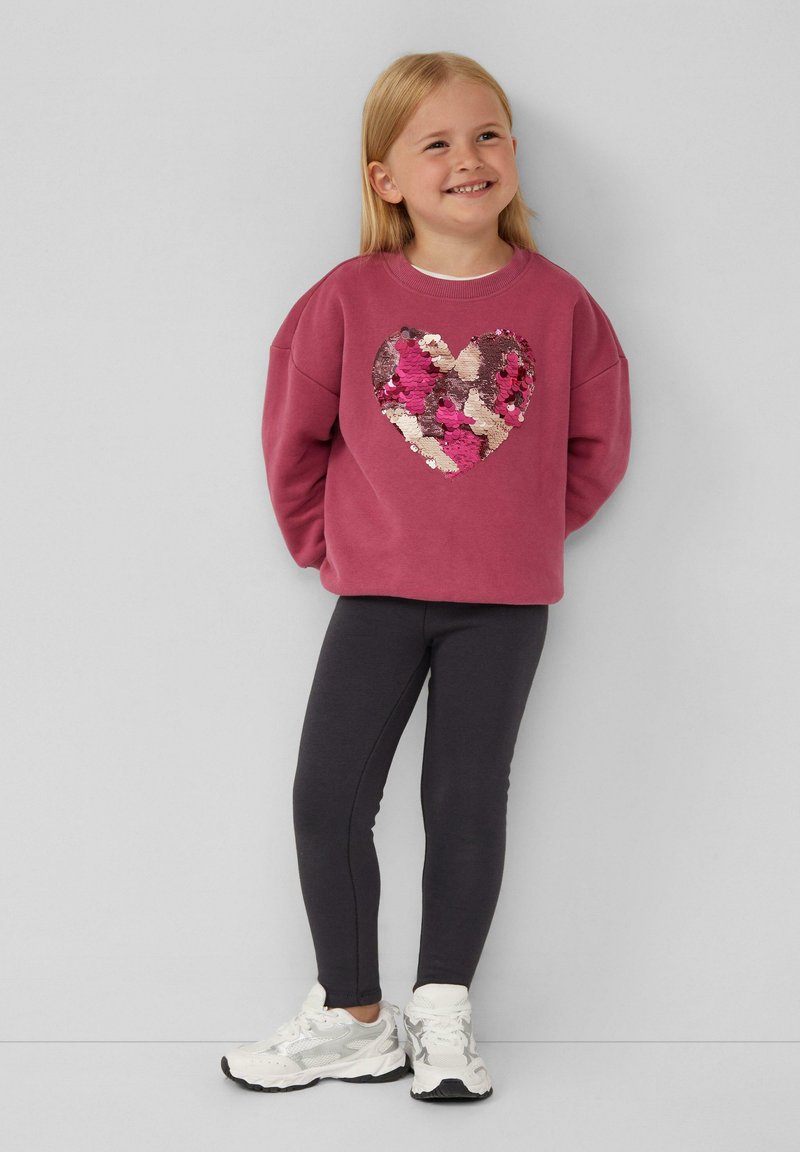 Rosa Sweatshirt mit einem großen, glitzernden Herzdesign, kombiniert mit dunkelgrauen Leggings und weißen Sneakers, auf einem hellen Hintergrund.