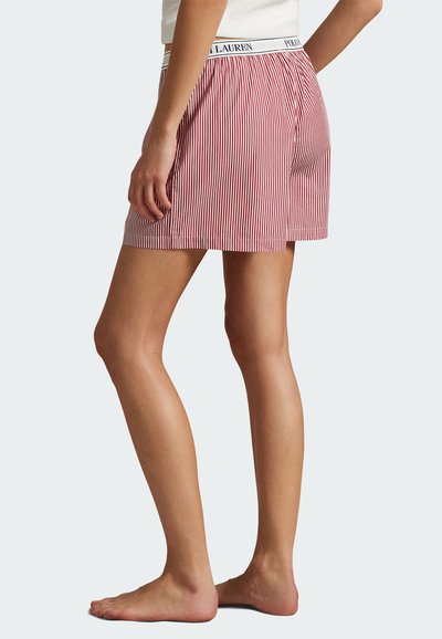 Kvinna som bär röda och vita vertikalt randiga shorts med ett vitt elastiskt midjeband med varumärke, stående barfotad på en ljus bakgrund.