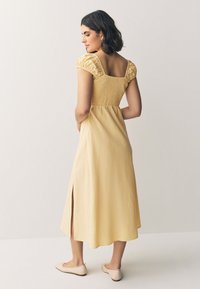 Abito midi giallo con maniche a palloncino, corpino arricciato e spacco laterale. Indossato con ballerine beige chiaro. Tessuto testurizzato con una finitura morbida.