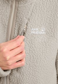 Veste en polaire gris clair avec une surface texturée, dotée d'une fermeture éclair à l'avant et d'un logo brodé sur la poitrine. La tirette de la fermeture éclair est en argent.