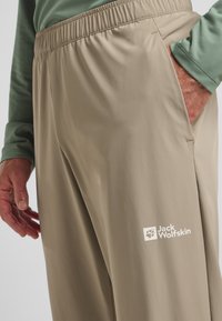 Pantaloni leggeri beige con elastico in vita, tasche laterali e logo Jack Wolfskin bianco sulla coscia sinistra. Texture liscia.