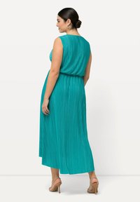 Vestido maxi sin mangas de color verde azulado, con pliegues y un ajuste relajado, cintura elástica. Textura suave, de longitud hasta el tobillo, combinado con sandalias de tacón desnudo.