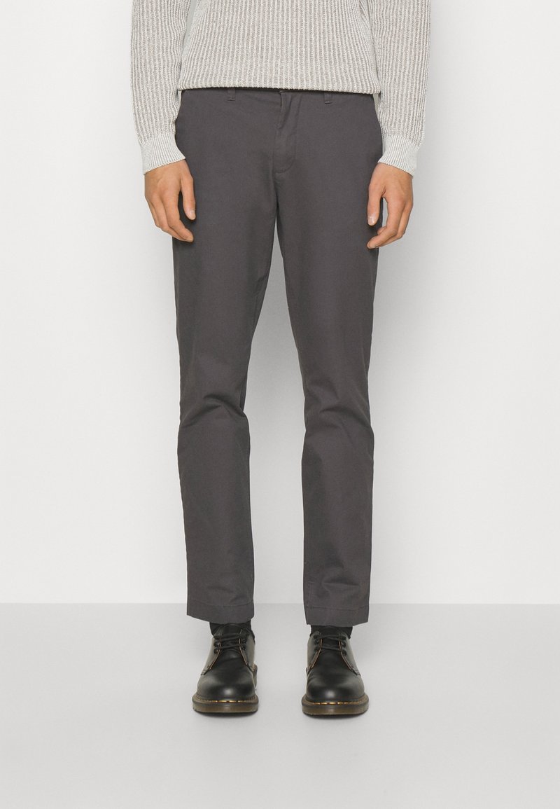 Homme portant un pantalon droit gris foncé, un pull en maille gris clair et des chaussures en cuir noires à lacets, debout devant un fond blanc uni.