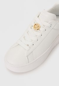 White leather sneaker with flat white laces, a textured rubber sole, and a gold circular logo accent on the side. Smooth surface, minimalist design.