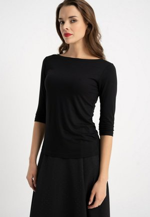 Kobieta o długich brązowych włosach i czerwonej szmince, ubrana w dopasowaną czarną bluzkę z długim rękawem oraz czarną spódnicę z subtelnym wzorem w kropki na białym tle.