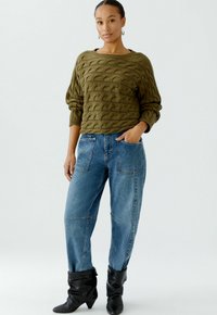 Olijfgroene gebreide trui met golfpatroon, gecombineerd met losse blauwe denimjeans en zwarte slouchy enkellaarzen. Model staat met de handen in de zakken.