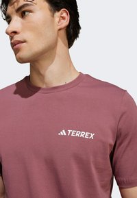 Bordová bavlněná tričko s kulatým výstřihem a krátkými rukávy, s bílým logem "TERREX" na levé části hrudi. Hladká textura, jednoduchý design.